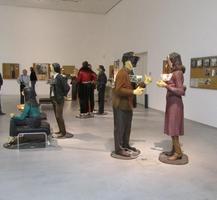 Lifelike sculptures of people standing around a room conversing; they have electronic or metal “faces” instead of real faces