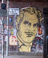 Large wall painting of man with moustache; text next to face says “It is hard to be a citizen.”