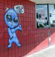 Blue alien advertising 2 hats for $25 on sign painted on wall of hat shop