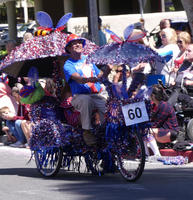 Man wearing bicycle with two large, gaudy umbrellas and tinsel on sides of wheels