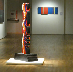 Colorful abstract statue in foreground; painting with vertical colored sstripe in background