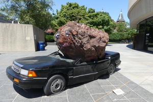 Large rock with eyes and mouth painted on it, crushing a car.