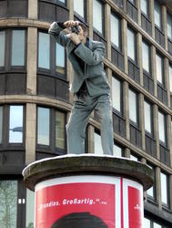 Statue of man with camera taking picture; statue is atop a cylindrical advert kiosk.