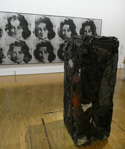 Rectangular sculpture made of crushed cars in foreground; Warhol’s pictures of Elizabeth Taylor in the background