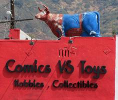 “Comics vs. Toys” store sign; on roof is a statue of a cow in a spiderman outfit.