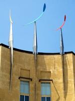 Three crescent-shaped sculptures (yellow, blue, and red) that rotate in wind, mounted on pointy pylons