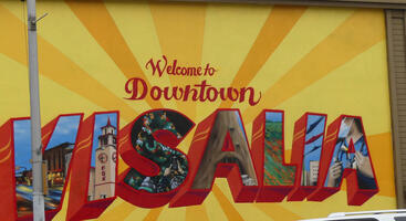 Mural in style of a postcard reading “Welcome to Downtown Visalia”, with local-themed scenes in the letters.