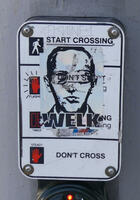 Traffic signal defaced with sticker of face of man labeled WELK
