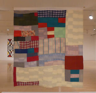 Large multi-colored quilt with plain and plaid pattterns, hanging from ceiling.