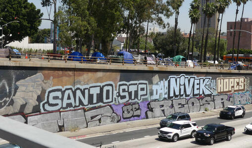 SANTO STP NIVEK HOPES painted on side of freeway wall; homeless encampment on surface street above wall.
