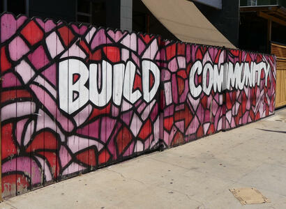 Fence with red geometric shapes in background; painted with words “Build Community”