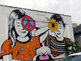 Boy and girl holding a heart; girl holds peace symbol over one eye and boy is holding sunflower over one eye.