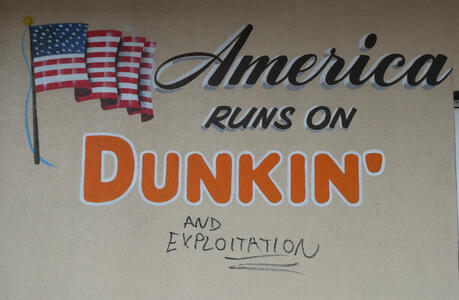 Sign: America runs on Dunkin’; underneath someone has written in marker “and exploitation”