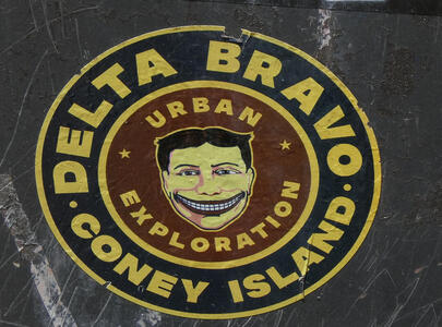 Widely grinning man; surrounding text in a circle: DELTA BRAVO CONEY ISLAND; inner circle text: URBAN EXPLORATION