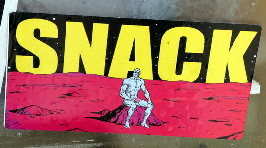 Picture of Dr. Manhattan (from “Watchmen” graphic novel) seated on a rock; text behind him reads SNACK in large yellow letters
