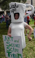 Woman wearing papier mache coffee up: “Wake up! Art and Science Scare Tyrants”