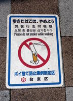 Sign on sidewalk reading “Please do not smoke while walking”; illustrated with a walking cigarette.
