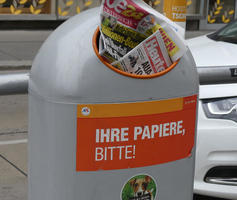 Trash can with orange sticker; can is filled with old newspapers.