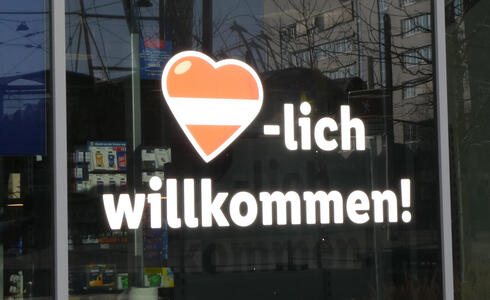 Sign with heart symbol, striped like Austrian flag, followed by words “-lich willkommen!”