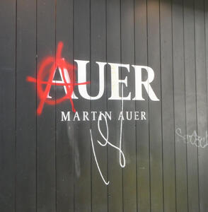 Sign for Martin Auer bakery; the A has been spray-painted to an anarchist symbol