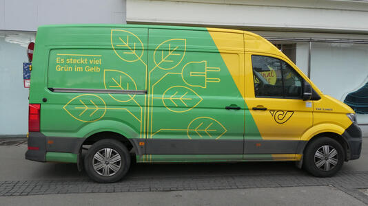 Side of a postal service truck; leaves on a tree, one of them an electric plug. Es steckt viel Grün im Gelb