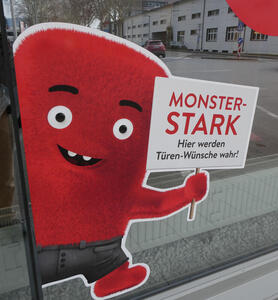 Cartoonish red monster holding sign: Monster-stark / Hier werden Türen-Wünsche wahr!