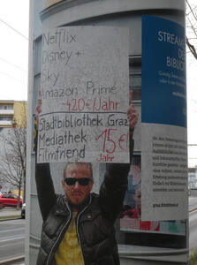 Man holding up a sign: Netflix Disney + Sky Amazon Prime: ~ 420€ / Jahr
Statdbibliothek Graz Mediathek Filmfriend: 15 € / Jahr