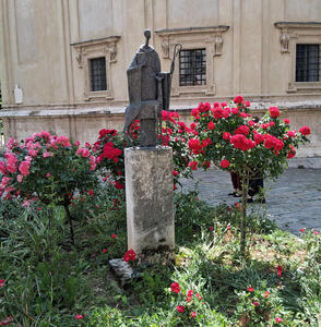 Abstract sculpture of man holding book and shepherd's crook. Sculpture is surrounded by roses.