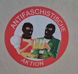 Picture of man and woman in typical Austrian garb wearing balaclavas. The text on the sticker reads „Antifaschistische Aktion“.