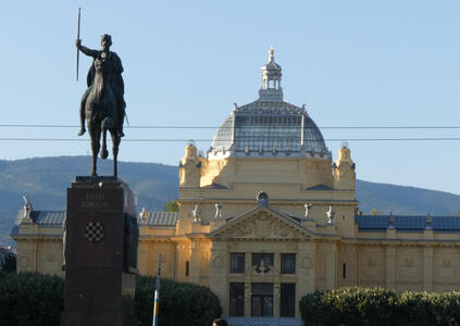 At left foreground, statue of a man on horseback, holding up a spear. At right in background, a large building with a dome and cupola.
