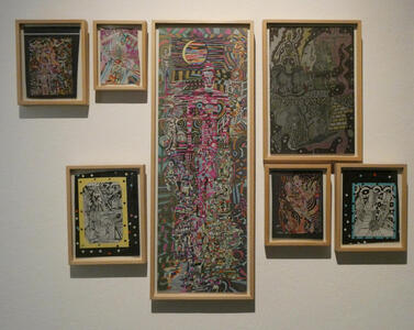 Wall with seven paintings in an asbtract style. The one in the middle has a recognizable figure of a human.