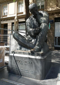 Statue of Nikola Tesla. He is sitting on the ground, one hand on his chin and the other on his knee.
