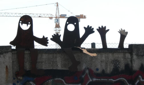Two metal ghost-like figures, one with vampire fangs and one with a wide-open mouth, and two hands, seated atop a wall.