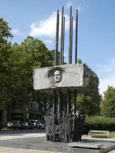 Sculpture of a man’s head emerging from a concrete block supported by steel rods. At the base of the rod are sculptures of people.