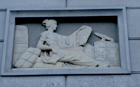 Carving of a reclining woman. Near her are bundles of goods.