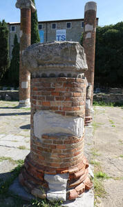 Brick cylinder with top of roman column on top of cylinder. Behind it are two brick columns.