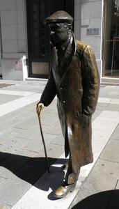 Statue of man with walking stick. He is wearing a long coat and an English driving cap