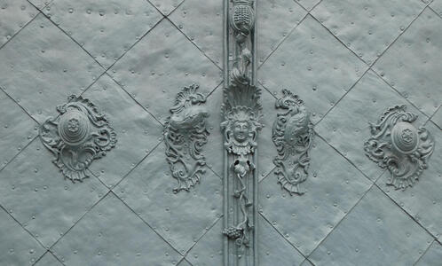 In center of door frame, a face. To either side, scrollwork that faintly resemles a sea lion; to its right, scrollwork on a half-sphere.  Below the face, a bunch of grapes.