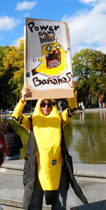 Woman in a banana costume holding a picture of a screaming banana with a crown. Text on sign reads “Power gone bananas”