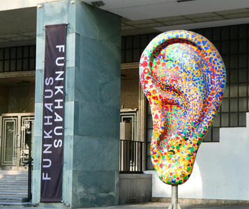 Large sculpture of a human ear, painted with splotches of varying colors. At left, a banner reading FUNKHAUS