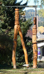At left, wood statue with frowning person who has two very long legs. At right, a totem pole-like wooden sculpture with a chess knight’s horse head