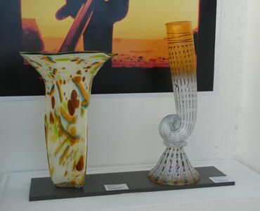 At left, a vase with spots of orange, red, green, and blue. At right, a vessel that curls into a spiral at the base. It is tinted orange at the top.