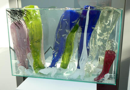 Abstract sculpture of what appear to be women’s legs wearing high heels. The legs are pink, green, blue, clear, and red.