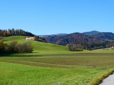 Open field in foreground, rolling hills and a small house in midground, and hills covered with trees in background.