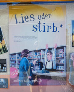 Woman in bandit mask pointing a banana at a librarian, with his hands above his head. The poster title reads „Lies oder stirb.“