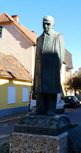 Man in long coat, looking downward pensively, hands behind back.