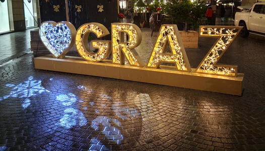 Lighted display; blue lights in a heart, followed by GRAZ with yellow lighting. Snowflakes are projected on the ground nearby.