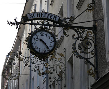 Clock at right angles to a building. Company name is Scheffer; clock is slightly tilted to the left.