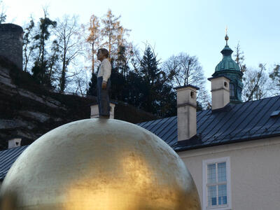 Large golden sphere with lifelike statue of person in white shirt, dress pants, and tie. Church spire in background at right.