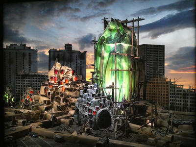 Painting of dilapidated building at left; at right, a glowing green large shape that is not quite a building.